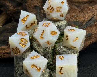 Op de afbeelding: Een set van zeven polyhedrale dobbelstenen in verschillende vormen, waaronder een D20, D12, D10, D8, D6 en D4. De dobbelstenen zijn wit met een marmer effect en goudkleurige cijfers. De dobbelstenen zijn gerangschikt op een bed van groen mos en hout.
