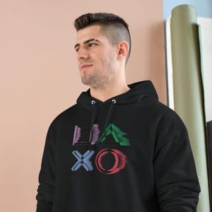 Puede incluir: Sudadera con capucha negra con un diseño gráfico colorido en la parte delantera. El diseño presenta las letras "LIA" en morado, un triángulo verde y las letras "XO" en azul y rojo. La sudadera tiene un bolsillo delantero y un logo de Champion en la manga.