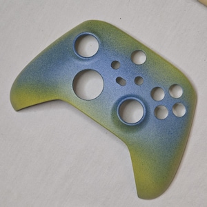May include: A custom-painted Xbox controller faceplate with a gradient design. The colour scheme transitions from yellow to blue, with a speckled texture. It features circular cutouts for buttons and joysticks.
