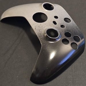 May include: A two-tone controller shell, changing from silver to black. The shell features multiple circular and oval button holes. The surface has a speckled texture and a glossy finish.