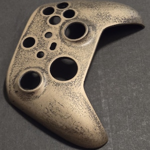 May include: A tan-colored video game controller shell with a distressed finish. The shell features multiple circular button holes and a curved ergonomic design. The controller has a worn, textured appearance, suggesting a vintage or custom aesthetic.