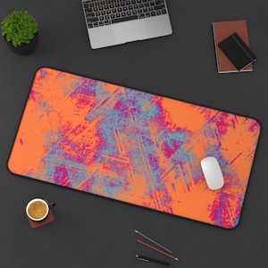 May include: An orange and blue abstract art mouse pad with a black border. A white computer mouse sits on the pad. A laptop, coffee cup, and stationery are also on the dark surface.