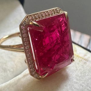 May include: A gold ring featuring a large, rectangular, vibrant pink gemstone. The gemstone is surrounded by a halo of small, clear stones. The ring's band is a delicate gold color, creating a luxurious and eye-catching piece of jewelry.