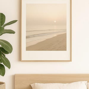 Könnte beinhalten: Ein gerahmter Druck einer Strandlandschaft. Das Bild zeigt einen Sandstrand, sanfte Wellen und einen sanften, goldenen Sonnenaufgang. Der Rahmen ist helles Holz, was eine beruhigende und ruhige Atmosphäre schafft.