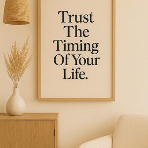 May include: A framed print with the text "Trust The Timing Of Your Life" in black lettering. The print is in a light wood frame and hangs on a cream-colored wall. A woven pendant light and a vase with dried plants are also visible.
