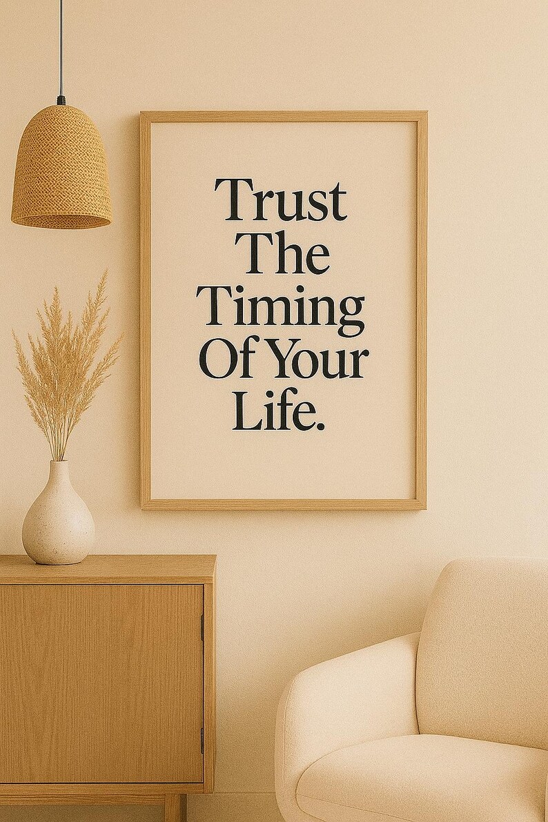 May include: A framed print with the text "Trust The Timing Of Your Life" in black lettering. The print is in a light wood frame and hangs on a cream-colored wall. A woven pendant light and a vase with dried plants are also visible.