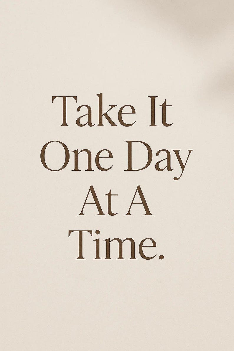 May include: A minimalist design featuring the text "Take It One Day At A Time" in a brown serif font. The text is centered on a beige background, creating a simple and motivational aesthetic.