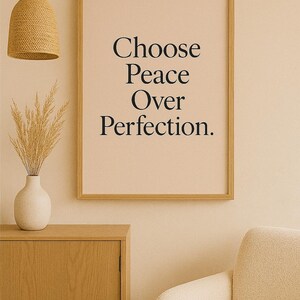 May include: A framed print with the text "Choose Peace Over Perfection" in a minimalist style. The artwork is displayed in a room with a neutral color palette, featuring a wooden cabinet, a beige armchair, and a woven pendant light.