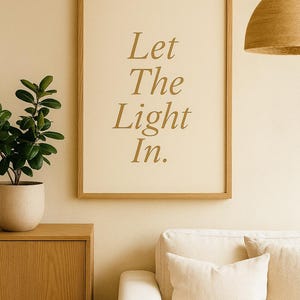 May include: A framed print with the words "Let The Light In" in gold lettering. The print is displayed on a beige wall above a cream-colored sofa. A wooden cabinet and a potted plant are to the left.