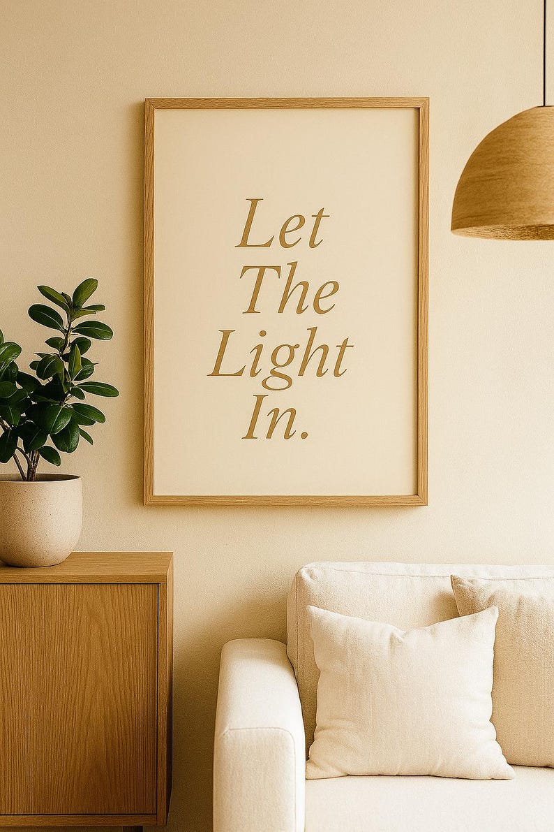 May include: A framed print with the words "Let The Light In" in gold lettering. The print is displayed on a beige wall above a cream-colored sofa. A wooden cabinet and a potted plant are to the left.