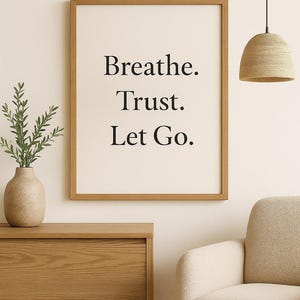 May include: A framed print with the words "Breathe. Trust. Let Go." in a minimalist black font. The print hangs above a wooden cabinet and next to a beige armchair. A woven pendant light and a vase with greenery are also visible.