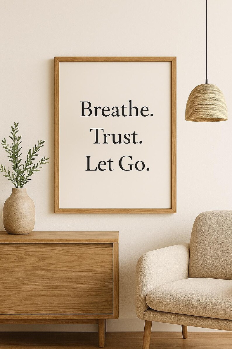May include: A framed print with the words "Breathe. Trust. Let Go." in a minimalist black font. The print hangs above a wooden cabinet and next to a beige armchair. A woven pendant light and a vase with greenery are also visible.