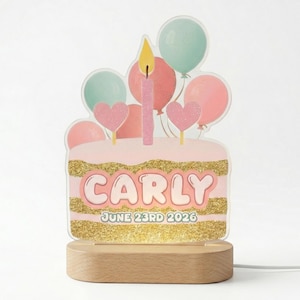May include: A clear acrylic birthday cake sign featuring a pink cake with gold glitter bands, a single pink candle, and pastel balloons. The text reads CARLY and JUNE 23RD 2026. The sign is mounted on a light-colored wooden base with a power cord.