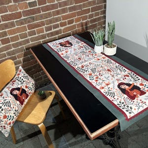 May include: A green and red table runner with a floral pattern and a woman's face in the center. The runner is on a black table with a wooden chair and a pillow with a similar pattern.