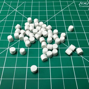 May include: A collection of small, white, cylindrical objects scattered across the frame. Each piece features a flat top and bottom with a smooth, solid white finish. The items are uniform in shape and size, appearing as individual components.