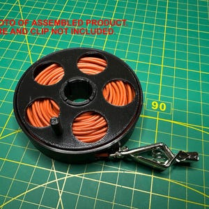 May include: A black spool filled with orange wire, with a silver clip attached. The spool has multiple circular openings and is set against a green grid background. Text at the top reads "PHOTO OF ASSEMBLED PRODUCT WIRE AND CLIP NOT INCLUDED."