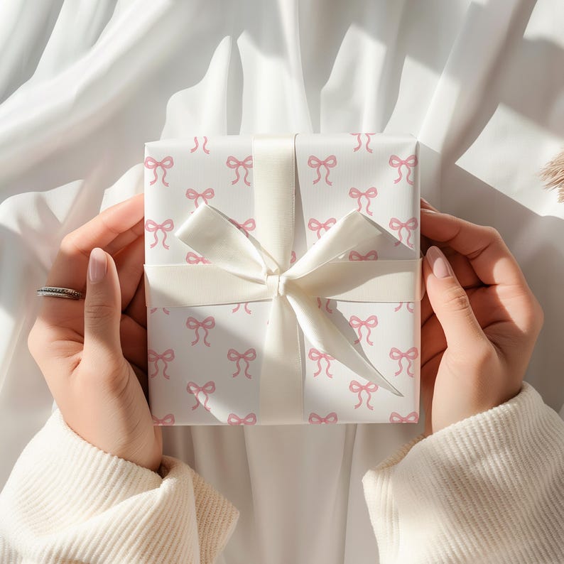 May include: A wrapped gift with white paper featuring a pattern of pink bows. The gift is tied with a wide, cream-colored ribbon and bow. The gift is held by two hands, with a ring on one finger.