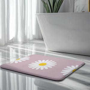 Könnte beinhalten: Rechteckige Badematte mit hellrosa Hintergrund und großen weißen Gänseblümchen-Blütenmustern mit gelben Zentren. Die Matte liegt vor einer weißen Badewanne in einem Badezimmer.