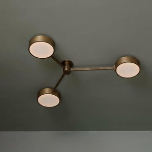 Peut inclure: Un plafonnier en bronze avec trois sources lumineuses circulaires. Les lumières sont disposées en triangle, chacune émettant une lueur douce et chaude. Le luminaire est fixé à un plafond gris foncé.