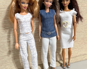May include: Three fashion dolls standing side by side. The first doll has blonde hair and wears a white top and pants with the word Together printed in script. The second doll has brown hair and wears a blue sleeveless top with a belt and white pants. The third doll has dark brown hair and wears a white dress with a heart-shaped embellishment.