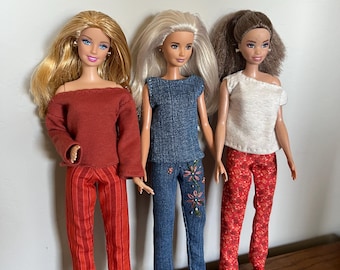 May include: Three dolls standing side by side. The left doll has blonde hair, a rust-colored long-sleeve top, and red striped pants. The center doll has platinum hair, a blue denim top, and blue jeans with floral embroidery. The right doll has brown hair, a white top, and red floral pants.