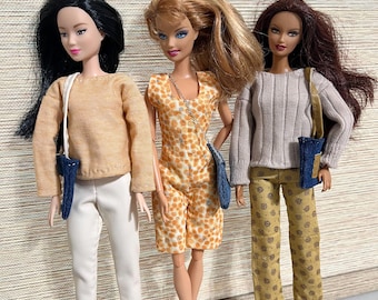 May include: Three fashion dolls are posed together, each wearing a unique outfit. One doll is in a tan sweater and white pants, another in an orange and white patterned romper, and the third in a gray sweater and yellow patterned pants. Each doll carries a small blue bag.