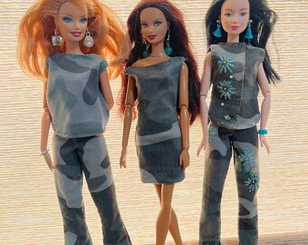 May include: Three dolls wearing matching gray and black camouflage outfits. The dolls have different skin tones and hairstyles. One doll wears a top and pants, another a dress, and the third a top and pants with floral accents. Each doll wears earrings and shoes.