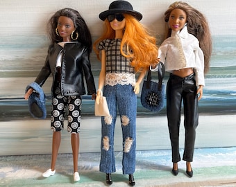 May include: Three fashion dolls standing side by side. The first wears a black jacket, patterned shorts, and white sneakers. The second has orange hair, a black hat, plaid top, and distressed jeans. The third wears a white shirt and black pants.