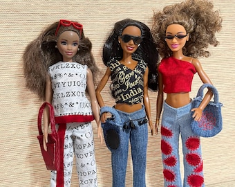 May include: Three fashion dolls standing side-by-side. The first wears a white top and pants with black letters and symbols, red stripes, and a red bag. The second wears a black graphic top, blue jeans, and black sneakers. The third wears a red top, blue jeans with red circular patches, and a blue bag.