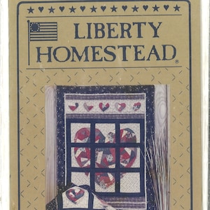 May include: A quilt pattern featuring a heart-shaped window with a red, white, and blue patchwork design. The pattern is titled "Liberty Homestead" and is from the "Pieced from the Heart Collection".