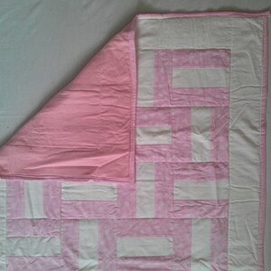 May include: A pink and white patchwork quilt with a geometric pattern. The quilt is folded in half, showing the pink backing.