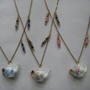 May include: Three gold chain necklaces with porcelain teapot pendants. Each teapot has a different floral design: blue flowers, white flowers, and pink roses. The chains have dangling charms: blue crystals, silver stars, and pink beads.