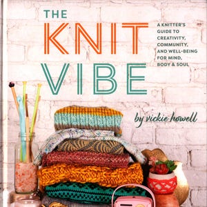 May include: The book "The Knit Vibe" is displayed with a stack of colorful knitted items. The cover text is orange and teal. Knitting needles are in a glass jar. A small pink radio and a succulent in a decorative pot are also visible.