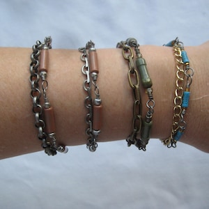 May include: Four bracelets stacked on a wrist. The bracelets are made of silver chain with brown and green beads.