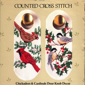 May include: Counted cross stitch kit for door knob decor featuring chickadees and cardinals. The kit includes two white door knob covers with embroidered birds, green foliage, and red berries. Finished size is 5" x 12".