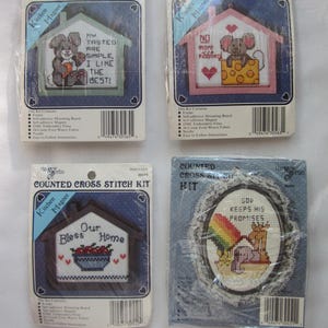 May include: Four counted cross stitch kits. Three are house-shaped kitchen magnets with stitched designs and text. One kit features a bear, another a mouse, and the third a bowl of fruit. The fourth kit is an oval with a rainbow and animals.