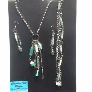 May include: A set of green and silver jewelry, including a necklace, earrings, and a bracelet. The necklace has a silver chain with a green glass bead and a charm made from recycled materials. The earrings and bracelet are also made with silver chains and green beads. The jewelry is handmade from reclaimed materials.