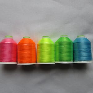 May include: Five spools of brightly colored thread are arranged in a row. The colors include pink, orange, yellow-green, green, and blue. Each spool has a white base and a green top.