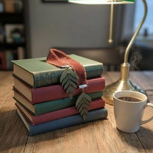 May include: A stack of vintage books with green, red, and blue covers, topped with a burgundy ribbon bookmark with leaf charms. A green desk lamp and a white mug of coffee sit nearby on a wooden surface.