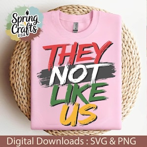 Op de afbeelding: Roze T-shirt met de tekst "They Not Like Us" in zwart, wit, rood, groen en geel gespatte letters.