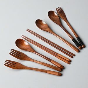 Puede incluir: Un conjunto de cubiertos de madera, que incluye cucharas, tenedores y palillos. Los utensilios son de madera marr&oacute;n c&aacute;lido, con algunos tenedores y palillos con detalles negros o envueltos. La disposici&oacute;n sugiere un tema de comedor o cocina.
