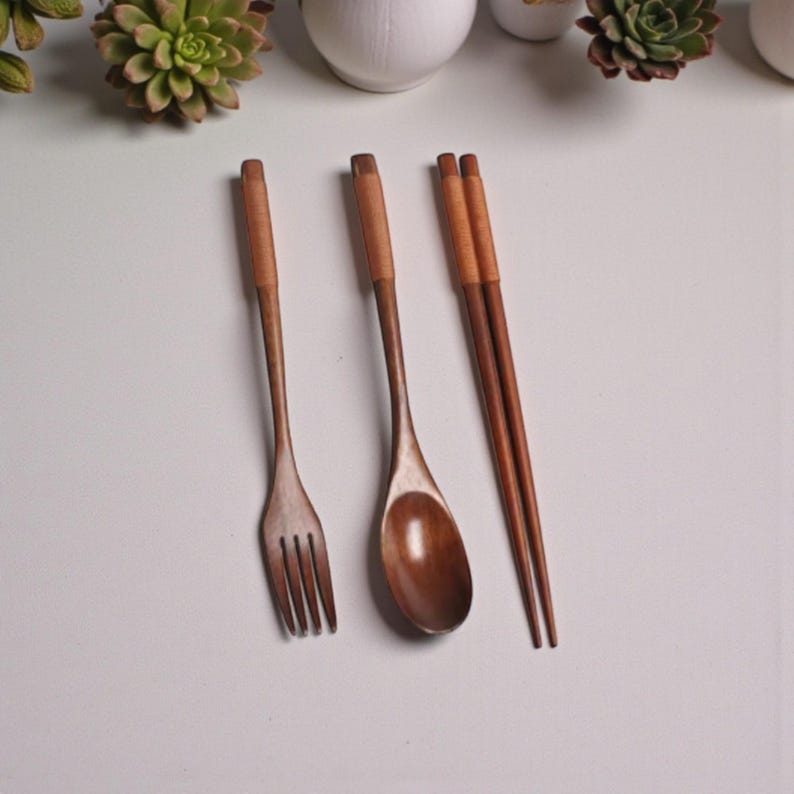 Puede incluir: Un juego de tenedor, cuchara y palillos de madera. Los utensilios est&aacute;n hechos de madera marr&oacute;n claro con un mango marr&oacute;n m&aacute;s oscuro. El tenedor tiene cuatro p&uacute;as y la cuchara tiene un cuenco redondeado. Los palillos son lisos y delgados.
