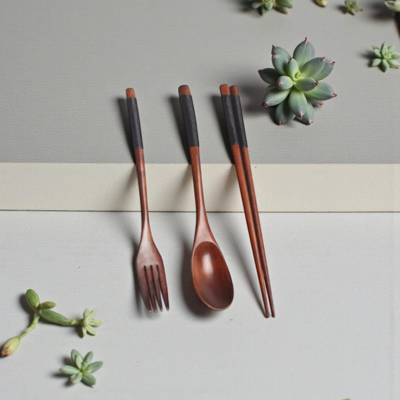 Puede incluir: Juego de cubiertos de madera con tenedor, cuchara y palillos. Los utensilios son de color marr&oacute;n c&aacute;lido con detalles negros. La cuchara tiene un cuenco redondeado y el tenedor tiene tres p&uacute;as. Los palillos est&aacute;n uno al lado del otro.