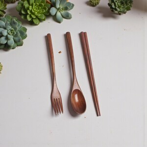 Puede incluir: Un juego de cubiertos de madera que incluye un tenedor, una cuchara y palillos chinos. Los utensilios son de madera marr&oacute;n oscuro y est&aacute;n colocados sobre una superficie blanca. El tenedor tiene cuatro p&uacute;as y la cuchara tiene un cuenco redondeado. Los palillos est&aacute;n paralelos.
