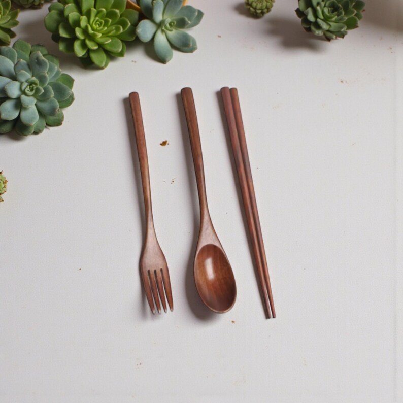 Puede incluir: Un juego de cubiertos de madera que incluye un tenedor, una cuchara y palillos chinos. Los utensilios son de madera marr&oacute;n oscuro y est&aacute;n colocados sobre una superficie blanca. El tenedor tiene cuatro p&uacute;as y la cuchara tiene un cuenco redondeado. Los palillos est&aacute;n paralelos.