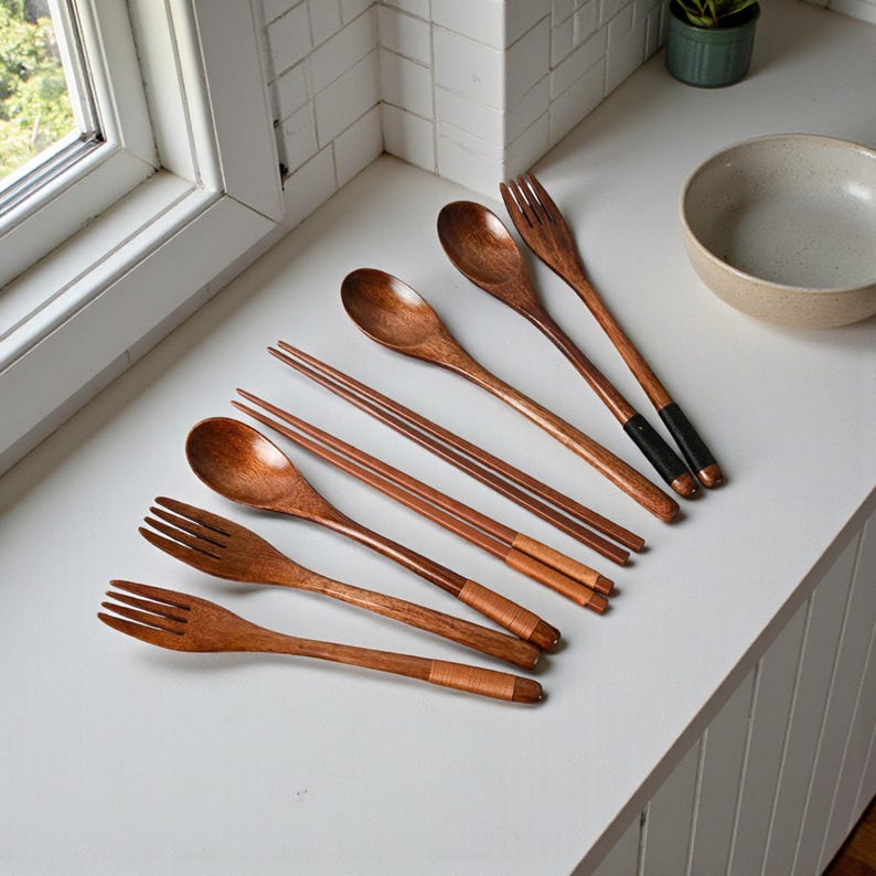 Puede incluir: Un juego de utensilios de madera, que incluye cucharas, tenedores y palillos, dispuestos sobre una superficie blanca. Los utensilios tienen un acabado de veta de madera natural con detalles de color cobre. Algunos tenedores tienen detalles negros. Tambi&eacute;n se ven un cuenco y una planta peque&ntilde;a.