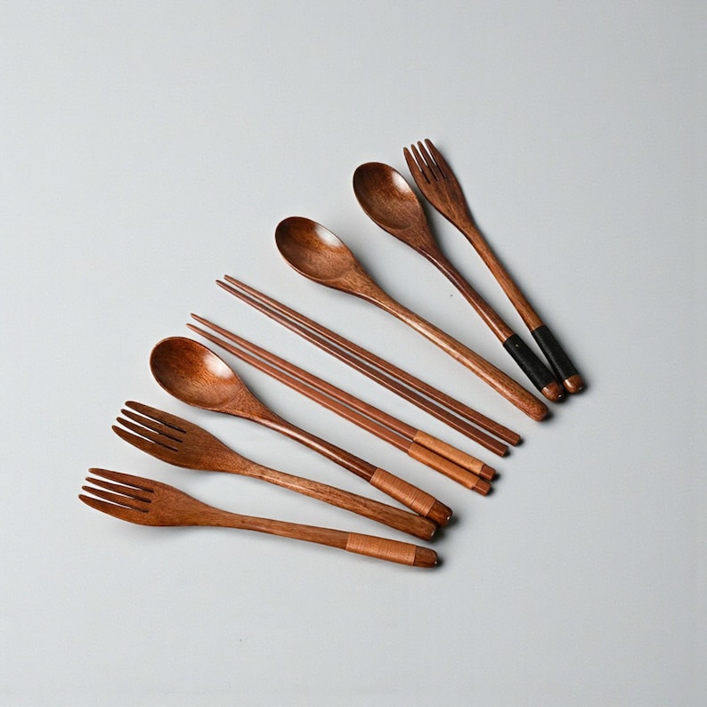 Puede incluir: Un conjunto de cubiertos de madera, que incluye cucharas, tenedores y palillos. Los utensilios son de madera marr&oacute;n c&aacute;lido, con algunos tenedores y palillos con detalles negros o envueltos. La disposici&oacute;n sugiere un tema de comedor o cocina.