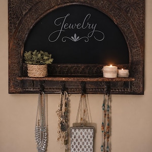 May include: A dark brown, carved wooden jewelry organiser with a chalkboard backing. The word "Jewelry" is written in cursive. The shelf holds a small plant, two candles, and several necklaces and a beaded purse hanging from hooks.