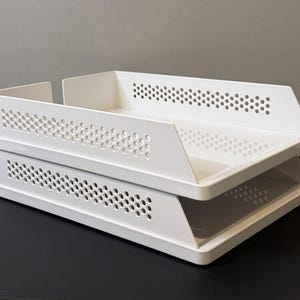Op de afbeelding: Een witte, tweelaagse bureau-organizer met een geperforeerd ontwerp. De trays zijn gestapeld, met schuine zijkanten en een rechthoekige vorm. De organizer is gemaakt van plastic en staat op een zwart oppervlak.