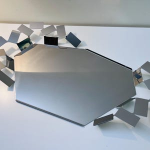 Könnte beinhalten: Ein moderner, geometrischer Wandspiegel mit einem großen, silbernen, vielseitigen zentralen Spiegel. Um den Spiegel herum befinden sich zahlreiche kleine, rechteckige, silbergraue Metallstücke, die ein einzigartiges, abstraktes Design bilden. Der Spiegel ist auf einer weißen Oberfläche montiert.